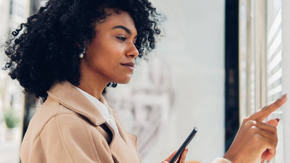 Side view of a confident adult woman using touch screen terminal on street while holding her phone.